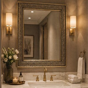 May include: A rectangular wall mirror with a wide, dark metallic frame featuring an intricate, repeating geometric pattern. The mirror is centered above a marble vanity countertop with a gold faucet and two matching wall-mounted light fixtures.