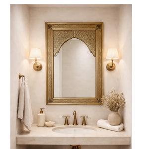 Salon avec un grand miroir doré orné au-dessus d'un canapé blanc. Des coussins beiges et une table basse ronde en bois avec des livres et des fleurs. Deux poufs en osier sont posés sur un tapis en jute.