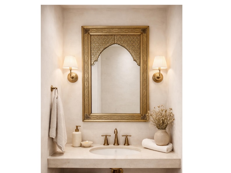 Salon avec un grand miroir doré orné au-dessus d'un canapé blanc. Des coussins beiges et une table basse ronde en bois avec des livres et des fleurs. Deux poufs en osier sont posés sur un tapis en jute.