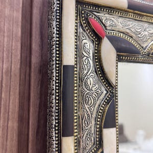 Pode incluir: Close-up de uma moldura de espelho decorativa. A moldura apresenta um design de filigrana de metal prateado com detalhes em preto, branco e vermelho. A moldura tem uma forma retangular com detalhes ornamentados e um acabamento polido.