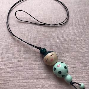 以下が含まれることがあります： 黒い紐のネックレスに木製のビーズのチャームが付いている。ビーズは顔を描いてあり、その下に緑と黒のドット柄のビーズが付いている。