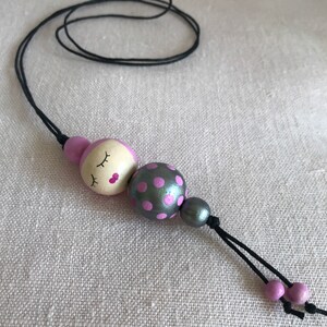 Pode incluir: Um colar de contas de madeira rosa e prata com um rosto dormindo em uma conta e bolinhas rosa em outra conta.