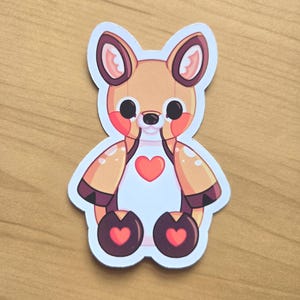 May include: A die-cut sticker featuring a stylized tan and white deer character with large black eyes, pink cheeks, and a white chest. The deer has a red heart on its chest and dark brown hooves, each decorated with a smaller red heart design.