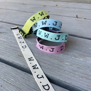 May include: Four woven bracelets in yellow, blue, light blue, and pink with the text "W.W.J.D." in black. A white woven bracelet with the same text is lying on a wooden surface.