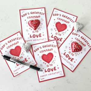 Op de afbeelding: Vijf witte kaarten met rode harten en de tekst "Gods grootste gebod is om lief te hebben" en "Gelukkig Valentijnsdag" erop gedrukt. Elke kaart heeft een klein chocoladehartje erop.