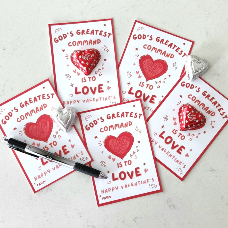 Op de afbeelding: Vijf witte kaarten met rode harten en de tekst "Gods grootste gebod is om lief te hebben" en "Gelukkig Valentijnsdag" erop gedrukt. Elke kaart heeft een klein chocoladehartje erop.