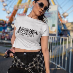 K&ouml;nnte beinhalten: Ein wei&szlig;es, bauchfreies T-Shirt mit einem schwarzen Barcode-Motiv und einem Rei&szlig;verschlussdetail. Das Shirt wird von einer Person mit dunklen Haaren und Sonnenbrille getragen. Die Person steht vor einem wei&szlig;en Zaun und einem Riesenrad.