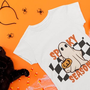 May include: White t-shirt with a black and white checkered pattern, a ghost holding a jack-o-lantern, and the text "Spooky Season!" in orange letters.