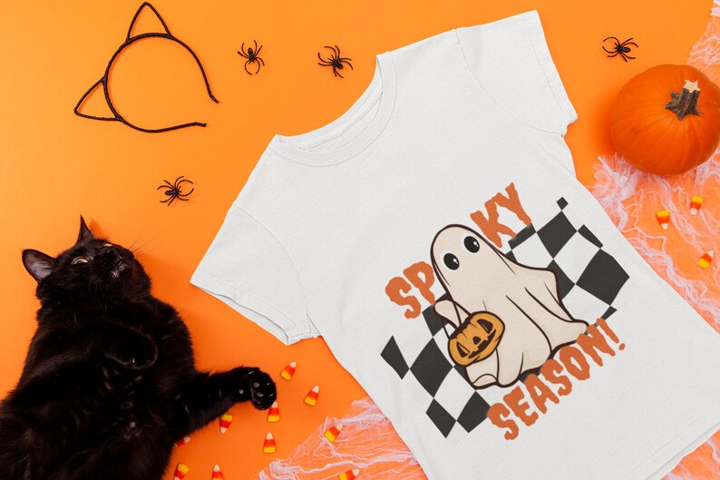 May include: White t-shirt with a black and white checkered pattern, a ghost holding a jack-o-lantern, and the text "Spooky Season!" in orange letters.
