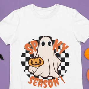 May include: White t-shirt with a graphic of a ghost holding a jack-o'-lantern in front of a checkered background. The text "Spooky Season!" is written below the ghost.