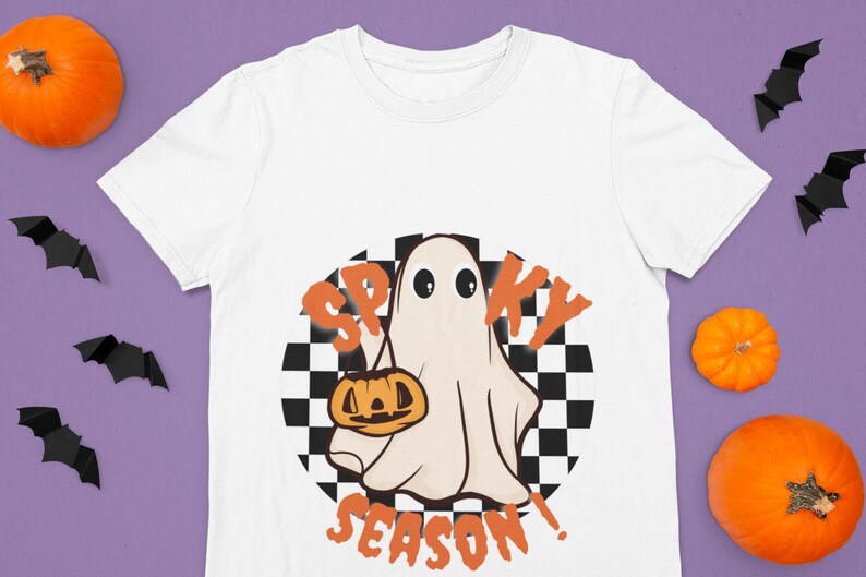 May include: White t-shirt with a graphic of a ghost holding a jack-o'-lantern in front of a checkered background. The text "Spooky Season!" is written below the ghost.