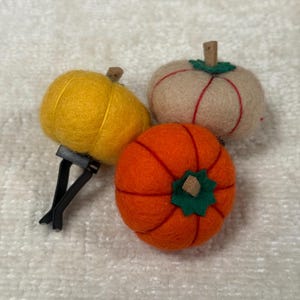 K&ouml;nnte beinhalten: Drei gefilzte K&uuml;rbis-Ornamente in Gelb, Beige und Orange. Jeder K&uuml;rbis hat einen kleinen braunen Stiel und ein gr&uuml;nes Filzblatt an der Oberseite. Der gelbe K&uuml;rbis ist an einem schwarzen Kunststoff-L&uuml;ftungsclip zur Verwendung im Auto befestigt.