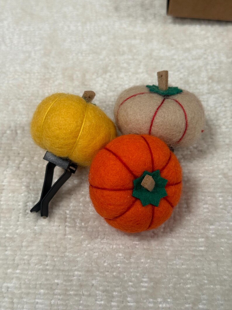 K&ouml;nnte beinhalten: Drei gefilzte K&uuml;rbis-Ornamente in Gelb, Beige und Orange. Jeder K&uuml;rbis hat einen kleinen braunen Stiel und ein gr&uuml;nes Filzblatt an der Oberseite. Der gelbe K&uuml;rbis ist an einem schwarzen Kunststoff-L&uuml;ftungsclip zur Verwendung im Auto befestigt.