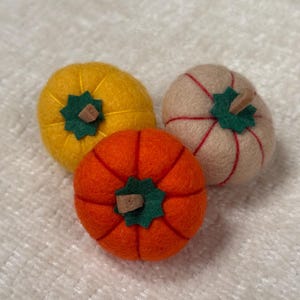 K&ouml;nnte beinhalten: Drei runde K&uuml;rbisse aus Filz in Gelb, Orange und Beige. Jeder K&uuml;rbis hat oben ein gr&uuml;nes, sternf&ouml;rmiges Filzblatt mit einem kleinen braunen Stiel. Die orangefarbenen und beigen K&uuml;rbisse haben rote N&auml;hte, der gelbe K&uuml;rbis hat gelbe N&auml;hte.