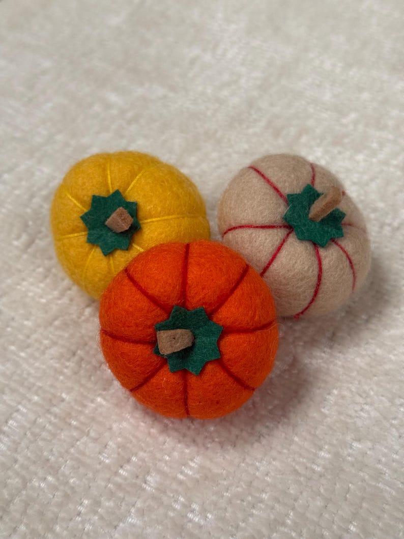 K&ouml;nnte beinhalten: Drei runde K&uuml;rbisse aus Filz in Gelb, Orange und Beige. Jeder K&uuml;rbis hat oben ein gr&uuml;nes, sternf&ouml;rmiges Filzblatt mit einem kleinen braunen Stiel. Die orangefarbenen und beigen K&uuml;rbisse haben rote N&auml;hte, der gelbe K&uuml;rbis hat gelbe N&auml;hte.