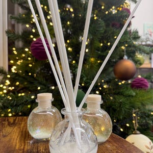 Peut inclure: Trois diffuseurs en verre avec des bâtonnets blancs. Les bouteilles contiennent un liquide clair et des paillettes. Les bouteilles ont des bouchons en bois. En arrière-plan, un sapin de Noël avec des décorations et des lumières.