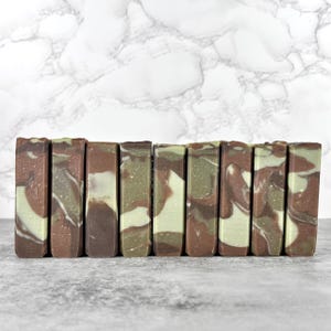 May include: A row of handmade soap bars with a marbled design. The bars feature a blend of brown, green, and white colors. The soaps are arranged side by side, showcasing their unique patterns and textures. The background is a marble pattern.