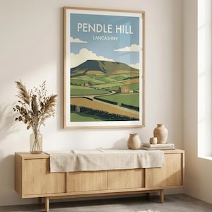 May include: A framed wall art print featuring a landscape illustration of Pendle Hill in Lancashire. The artwork displays a green hilly terrain, a winding road, stone walls, and a small house under a blue sky with white clouds. The text reads PENDLE HILL LANCASHIRE.