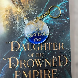 Puede incluir: Un llavero de resina azul y blanco con el texto "No rope can hold me" en una cadena dorada. El llavero está encima de un libro titulado "Daughter of the Drowned Empire" de Frankie Diane Mallis.