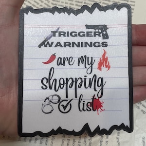 May include: A white sticker with black text that reads "Trigger Warnings are my shopping list". The sticker has a red chili pepper, a gun, a flame, handcuffs, and a check mark. The sticker has a white background with black lines.