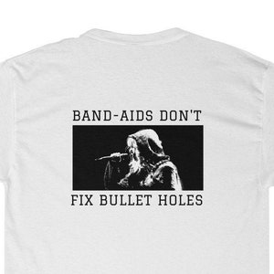 Könnte beinhalten: Weißes T-Shirt mit einer schwarz-weißen Grafik auf dem Rücken. Die Grafik zeigt eine Person, die in ein Mikrofon singt, mit dem Text "BAND-AIDS DON'T FIX BULLET HOLES" über und unter dem Bild.