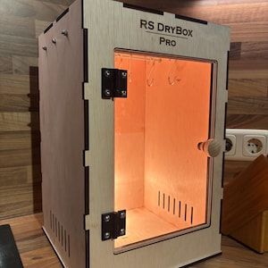 May include: A wooden box with the text "RS DRYBOX PRO" on the top. The box has a clear glass door, metal hinges, and a wooden handle. Inside, there are metal hooks and a warm orange light.