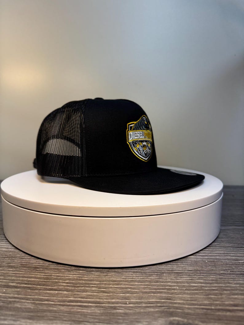 Pode incluir: Bon&eacute; de caminhoneiro preto com parte traseira em tela e aba reta. A frente do bon&eacute; apresenta um emblema em forma de escudo com a palavra "DIESEL" em letras douradas. O emblema tamb&eacute;m inclui detalhes em amarelo e cinza.