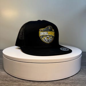 Pode incluir: Bon&eacute; de caminhoneiro preto com parte traseira em malha e aba reta. A frente do bon&eacute; apresenta um emblema em forma de escudo com as palavras "DIESEL POWER" em amarelo e branco, juntamente com um design gr&aacute;fico.