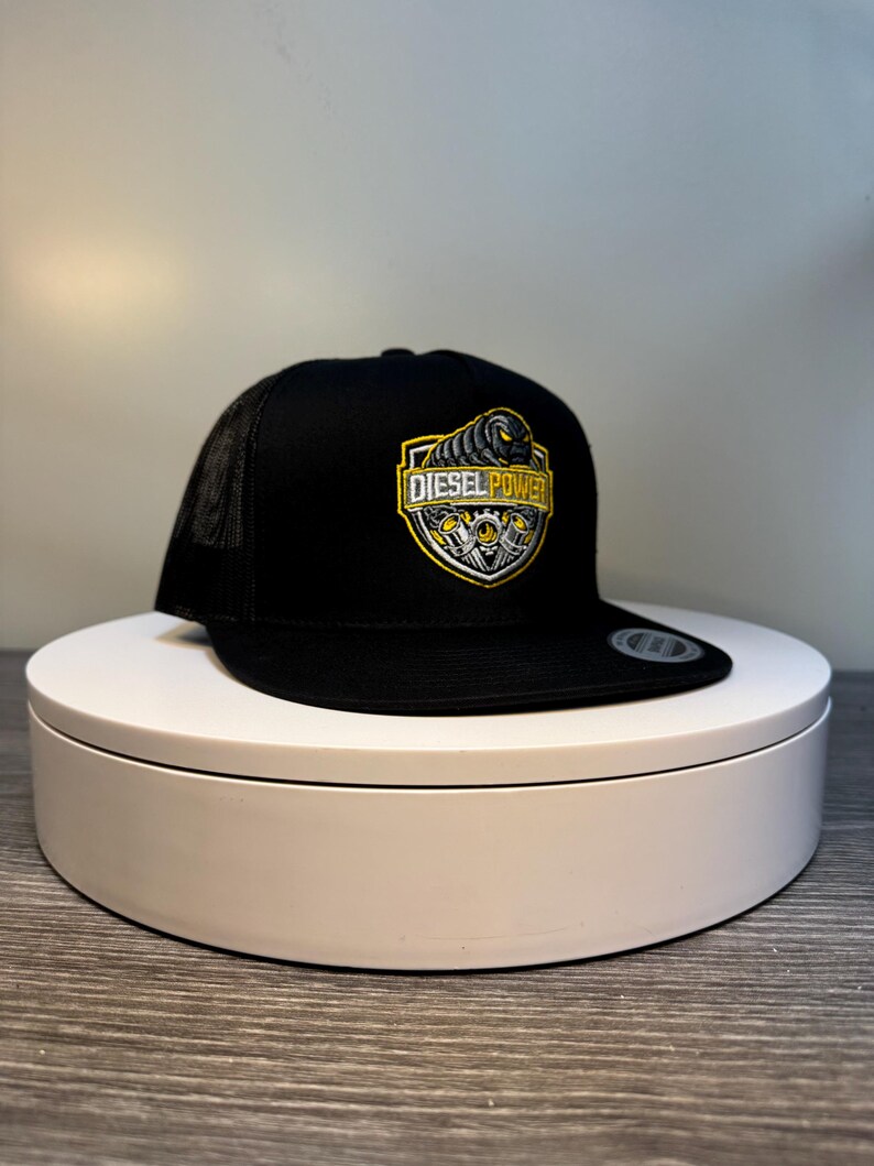 Pode incluir: Bon&eacute; de caminhoneiro preto com parte traseira em malha e aba reta. A frente do bon&eacute; apresenta um emblema em forma de escudo com as palavras "DIESEL POWER" em amarelo e branco, juntamente com um design gr&aacute;fico.