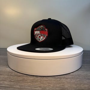 Pode incluir: Bon&eacute; de caminhoneiro preto com parte traseira em tela e aba reta. A frente apresenta um emblema vermelho, branco e preto com as palavras "DIESEL POWER". O bon&eacute; est&aacute; em um suporte circular branco.