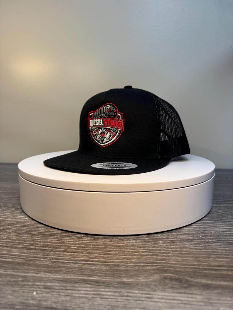 Pode incluir: Bon&eacute; de caminhoneiro preto com parte traseira em tela e aba reta. A frente apresenta um emblema vermelho, branco e preto com as palavras "DIESEL POWER". O bon&eacute; est&aacute; em um suporte circular branco.