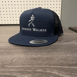 May include: A navy blue trucker hat with a white embroidered Johnnie Walker logo and a black mesh back. The hat has a snap closure and a silver label that reads "The Authentic Snapback Yupoong Since 1974".