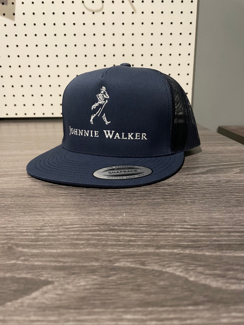May include: A navy blue trucker hat with a white embroidered Johnnie Walker logo and a black mesh back. The hat has a snap closure and a silver label that reads "The Authentic Snapback Yupoong Since 1974".
