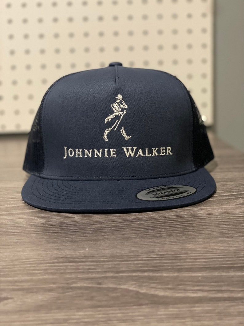 May include: A navy blue trucker hat with a white mesh back. The hat has a white embroidered logo of a walking man and the text "JOHNNIE WALKER".