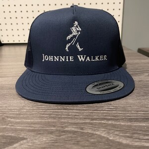 May include: A navy blue trucker hat with a white embroidered Johnnie Walker logo and a walking man figure. The hat has a black mesh back and a white "Snapback" label.