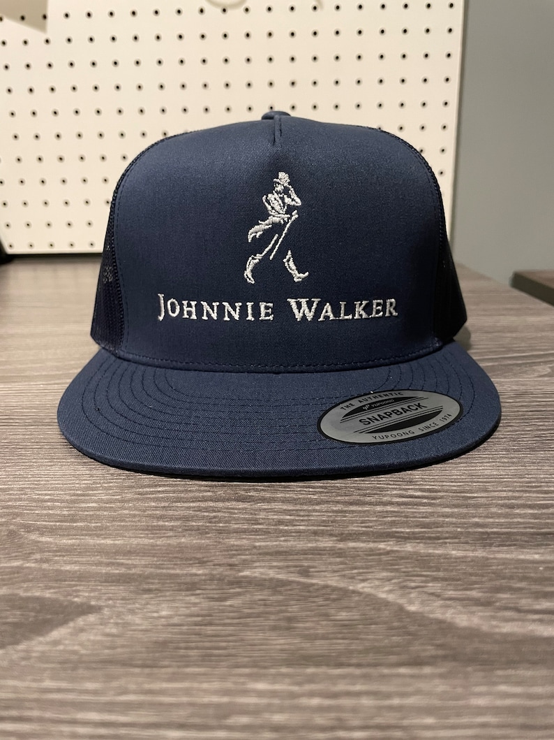May include: A navy blue trucker hat with a white embroidered Johnnie Walker logo and a walking man figure. The hat has a black mesh back and a white "Snapback" label.