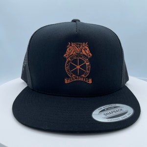 May include: Black trucker hat with a brown embroidered logo featuring two horses, a wheel, and the text "International Brotherhood of Teamsters". The hat has a black mesh back and a black snap closure.