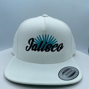 May include: White baseball cap with a blue and black embroidered logo that says "Jalisco". The cap has a black "The Authentic" sticker on the side.