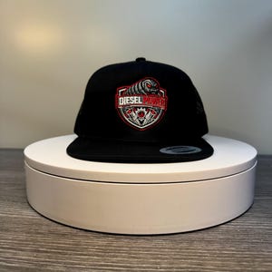 Pode incluir: Bon&eacute; snapback preto com o logotipo Diesel Power. O logotipo apresenta um design de escudo vermelho e branco com o texto "DIESEL POWER" e um gr&aacute;fico de um motor. O bon&eacute; &eacute; exibido em um suporte circular branco.
