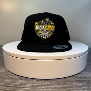 Pode incluir: Bon&eacute; de caminhoneiro preto com parte traseira em malha e aba reta. A frente apresenta um patch bordado amarelo e preto com "DIESEL POWER" e um gr&aacute;fico de motor.