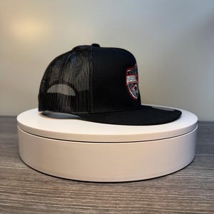 Pode incluir: Bon&eacute; de caminhoneiro preto com parte traseira em tela e aba reta. O painel frontal apresenta um emblema em forma de escudo com a palavra "DIESEL" em um design vermelho e prateado. O bon&eacute; &eacute; exibido em uma plataforma girat&oacute;ria branca.