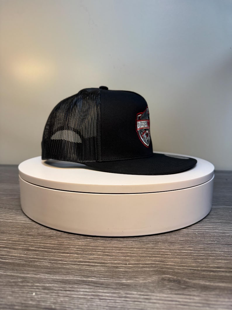 Pode incluir: Bon&eacute; de caminhoneiro preto com parte traseira em tela e aba reta. O painel frontal apresenta um emblema em forma de escudo com a palavra "DIESEL" em um design vermelho e prateado. O bon&eacute; &eacute; exibido em uma plataforma girat&oacute;ria branca.