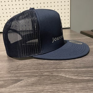 May include: A navy blue trucker hat with a mesh back and a white embroidered logo that says "JOHNNIE".