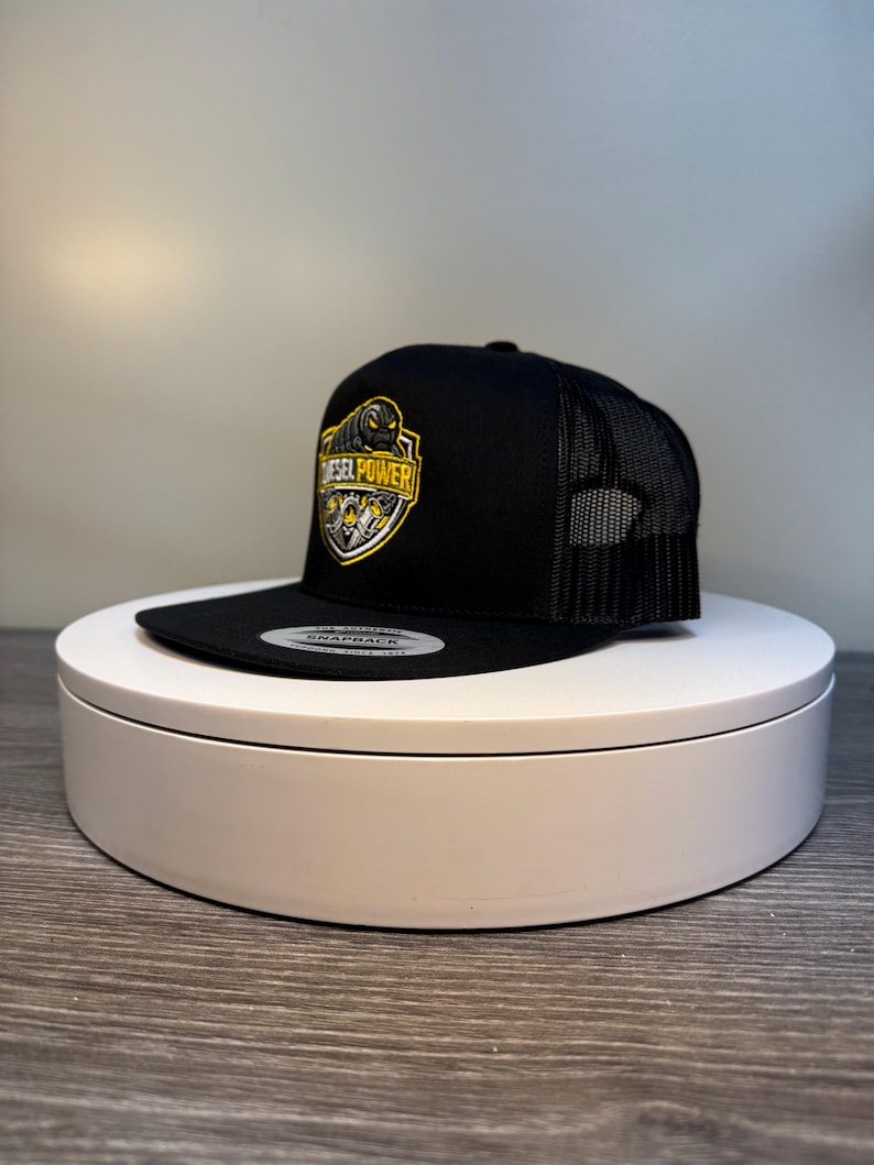 Pode incluir: Bon&eacute; snapback preto com parte traseira em rede e aba reta. A frente apresenta um emblema amarelo e preto com as palavras "DIESEL POWER". O bon&eacute; &eacute; exibido em um suporte circular branco.