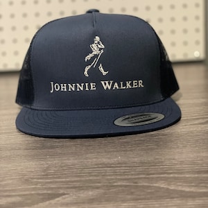 May include: A navy blue trucker hat with a white mesh back. The hat features a white embroidered Johnnie Walker logo with a walking man figure.