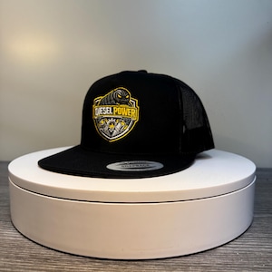 Pode incluir: Bon&eacute; de caminhoneiro preto com aba curva e parte traseira em tela. A frente apresenta um emblema amarelo e preto com as palavras "DIESEL POWER" e um gr&aacute;fico de pist&atilde;o. O bon&eacute; est&aacute; em um suporte girat&oacute;rio branco.