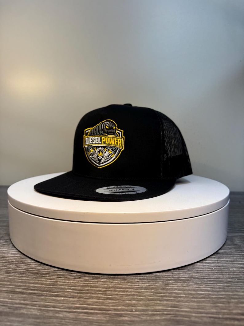 Pode incluir: Bon&eacute; de caminhoneiro preto com aba curva e parte traseira em tela. A frente apresenta um emblema amarelo e preto com as palavras "DIESEL POWER" e um gr&aacute;fico de pist&atilde;o. O bon&eacute; est&aacute; em um suporte girat&oacute;rio branco.