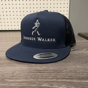 May include: A navy blue trucker hat with a white embroidered Johnnie Walker logo and a black mesh back. The hat has a white sticker that says "The Authentic Yupoong Snapback Since 1974."