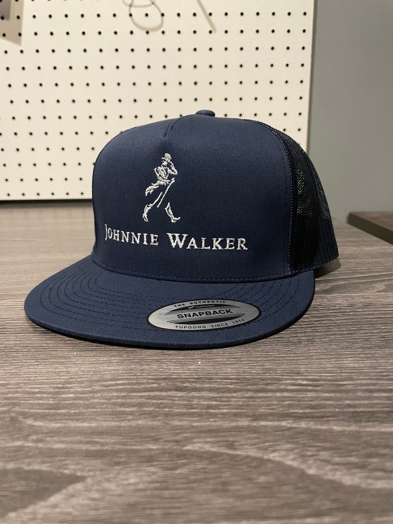 May include: A navy blue trucker hat with a white embroidered Johnnie Walker logo and a black mesh back. The hat has a white sticker that says "The Authentic Yupoong Snapback Since 1974."