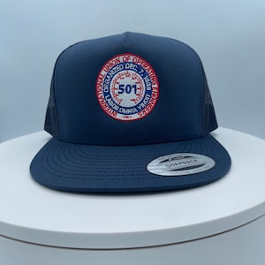 May include: A navy blue trucker hat with a black mesh back. The front of the hat features a red, white, and blue embroidered patch with the text "International Union of Operating Engineers Organized Dec. 7, 1896 Labor Omnia Vincit" and the number "501".